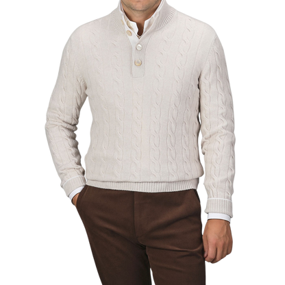 A man is wearing a cream Pure Cashmere Quarter Button Sweater by Gran Sasso over a white button-up shirt, paired with brown trousers. The top button of the cable-knitted sweater is undone. The background is plain gray.