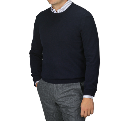 Clad in a Gran Sasso Navy Blue Cashmere Crew Neck Sweater and striped shirt, paired with gray pants, a person stands against a plain backdrop. This sustainable knitwear ensemble blends style and responsibility seamlessly, though their face remains unseen.