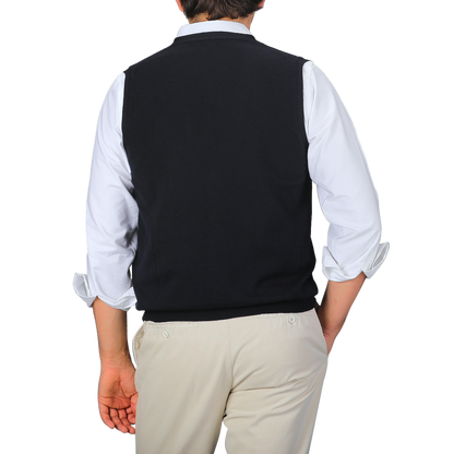 A person stands facing away, layering a white long-sleeve shirt with rolled-up sleeves and the Gran Sasso Navy Blue Extra Fine Merino Wool Vest over beige pants against a gray background.