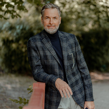 A middle-aged man with gray hair and a beard stands outdoors by a red railing, wearing a Gran Sasso Navy Blue Extra Fine Merino Wool Rollneck under a slim fit plaid blazer and light pants. Trees fill the background.