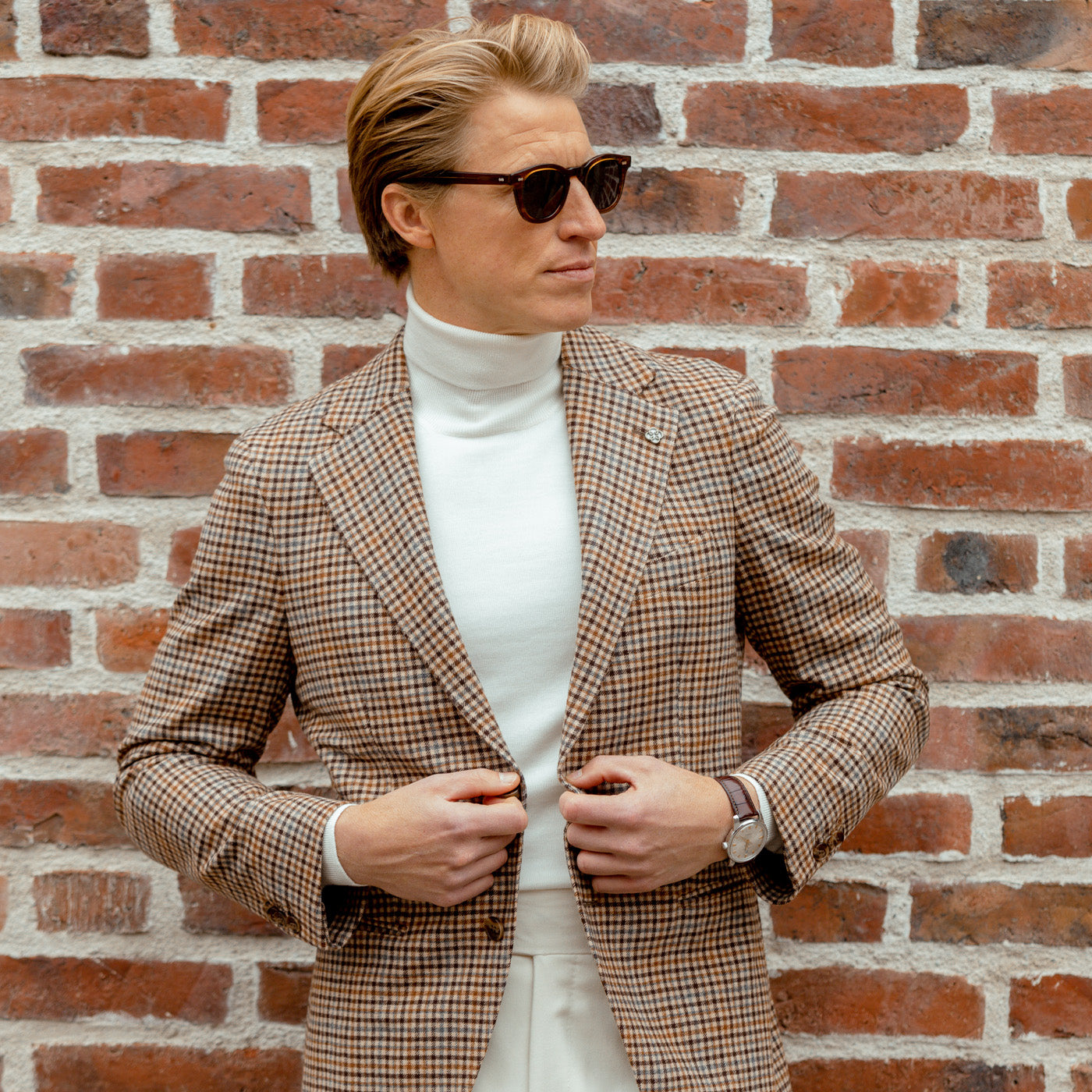 A person in sunglasses and a checked blazer leans against a brick wall, casually adjusting their Off White Extra Fine Merino Rollneck for that signature touch of Gran Sasso elegance.