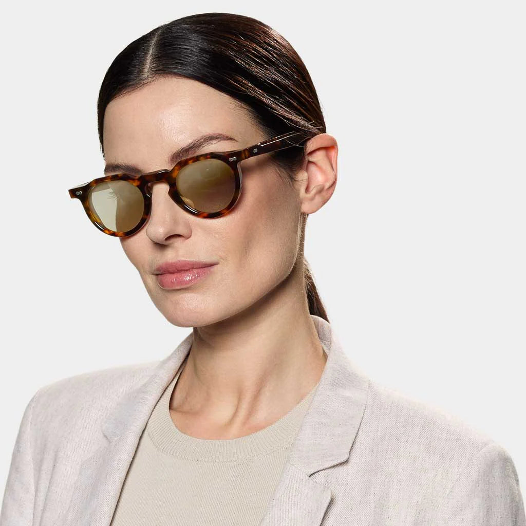A woman with straight dark hair in a low ponytail wears TBD Eyewear's Hem Eco Spotted Havana sunglasses with Mirror Green lenses, paired with a light beige blazer over a cream top, against a plain light background. , available at Richard Gelding Mayfair