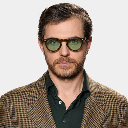A man with short brown hair and a beard wears TBD Eyewear's Hem River | Light Green glasses. He looks serious in a brown houndstooth blazer over a dark green shirt, standing against a plain white background. , available at Richard Gelding Mayfair