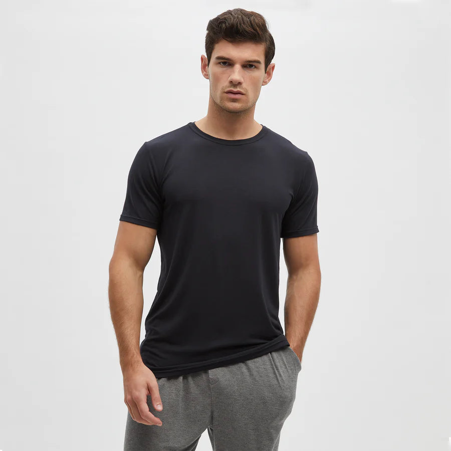 A man with short dark hair models the Homebody Black Modal Classic Short Sleeve T-Shirt and gray jogger pants against a light gray background, standing with his left hand in his pocket and a neutral expression, looking at the camera. , available at Richard Gelding Mayfair