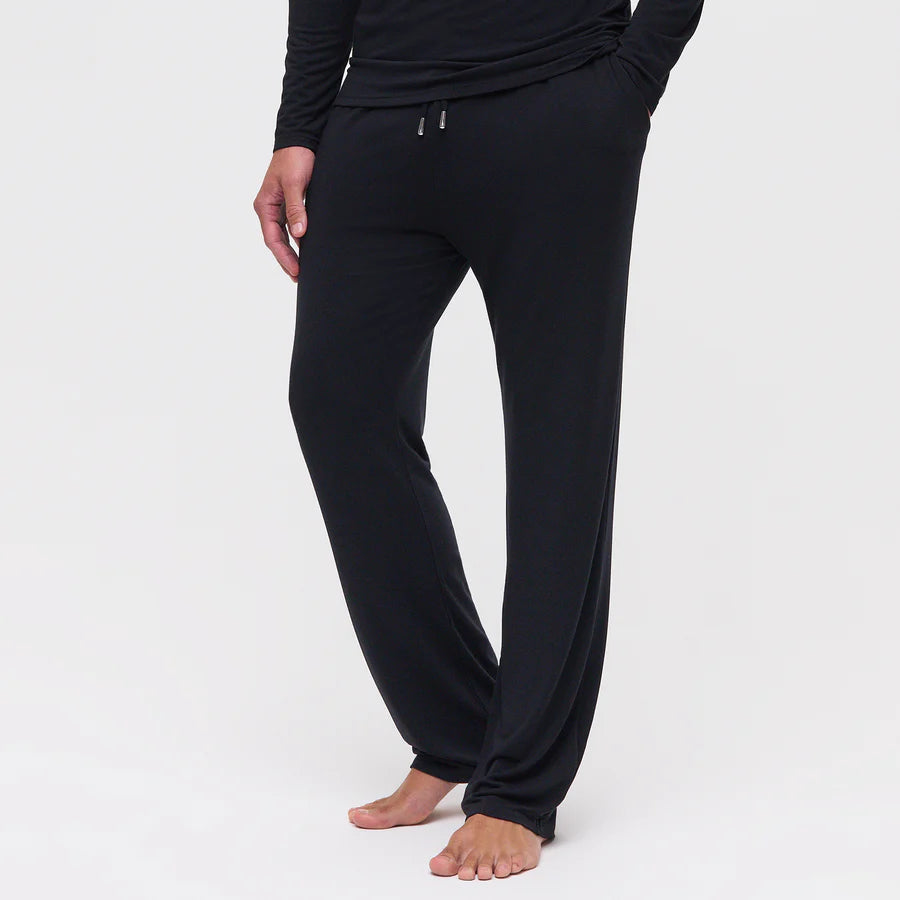 A person stands barefoot against a white background, displaying the relaxed fit and soft drape of Homebody’s Black Modal Modern Pyjama Trousers with a drawstring waist—ideal for those seeking comfortable men’s sleepwear. , available at Richard Gelding Mayfair