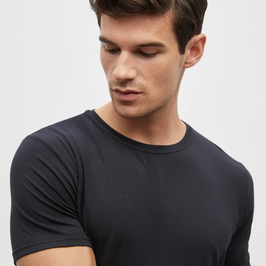 A man with short brown hair wears a fitted black Homebody Modal Classic Short Sleeve T-Shirt, looking down to his left with a neutral expression. The simple background highlights him and this versatile essential. , available at Richard Gelding Mayfair