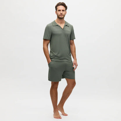 A man stands barefoot against a plain white background, wearing a Charred Green Collared Modal Marlow Polo T-shirt and matching shorts made from breathable fabric, with his hands in his pockets.