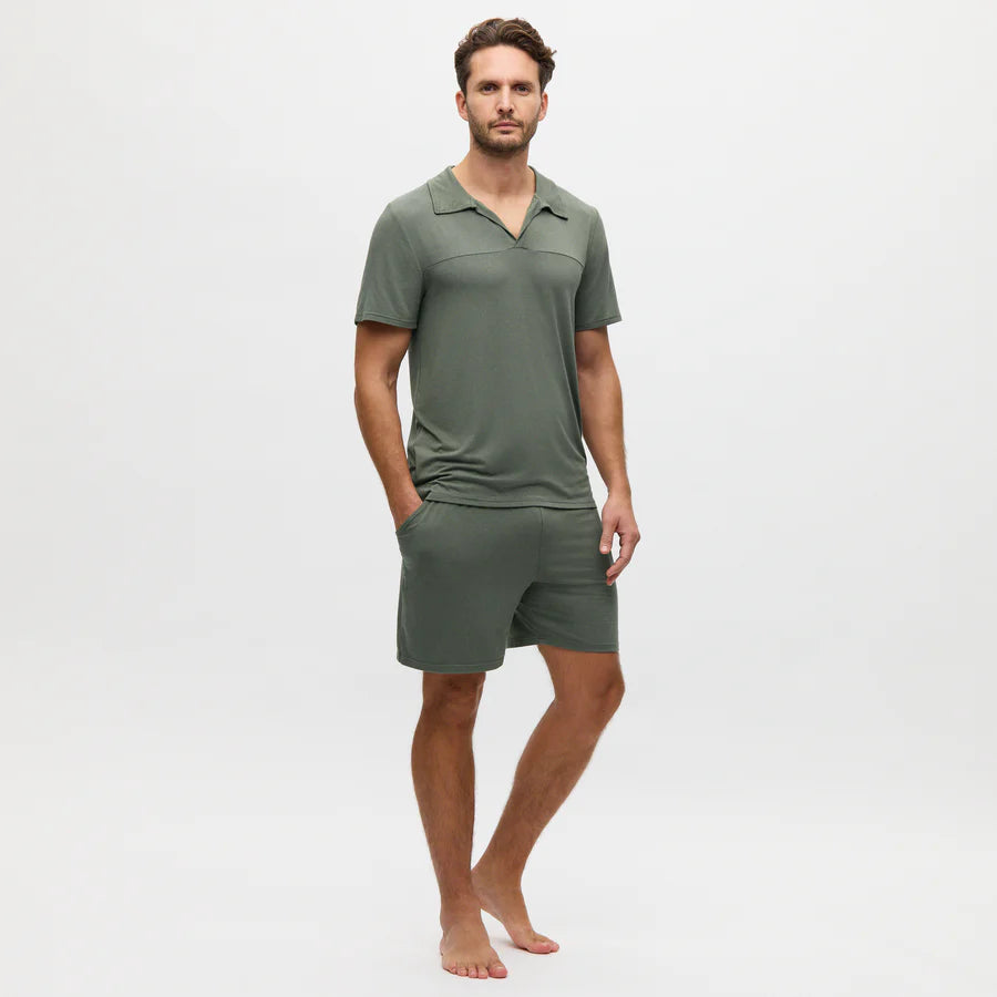 A man stands barefoot against a plain white background, wearing a Charred Green Collared Modal Marlow Polo T-shirt and matching shorts made from breathable fabric, with his hands in his pockets.