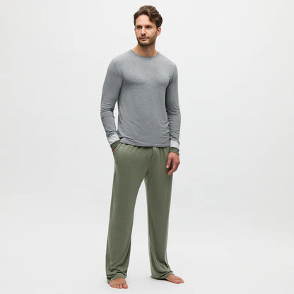 A man stands barefoot against a plain white background, wearing a light gray long-sleeve shirt and Homebody’s Charred Green Modal Duo Trousers. The trousers are loose-fitting, made from breathable fabric. He faces slightly left with a neutral expression, left hand in pocket. , available at Richard Gelding Mayfair