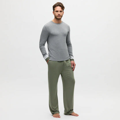 A man with short brown hair and a trimmed beard stands barefoot against a white background, wearing a fitted gray long-sleeve shirt and Homebody's Charred Green Modal Duo Trousers, his hands resting in the pants pockets. , available at Richard Gelding Mayfair