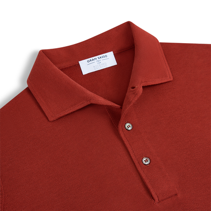 A close-up of the Rust Red Silk Pique Polo by Gran Sasso, featuring a textured knit, classic collar, two beige buttons, and an inner white “Gran Sasso” label, neatly laid flat on a white background. , available at Richard Gelding Mayfair