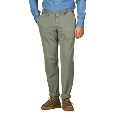 A person wearing a blue shirt, Green Washed Cotton Gabardine Slim Chinos by Hiltl, and brown shoes stands with hands in pockets.
