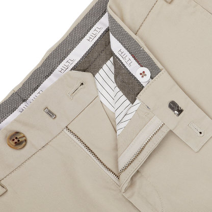 Close-up of khaki beige chinos with an unzipped fly revealing the interior lining; features include belt loops, buttons, and a waistband showcasing the "Hiltl" brand label. These regular fit pants are made from pure cotton twill for both comfort and style.