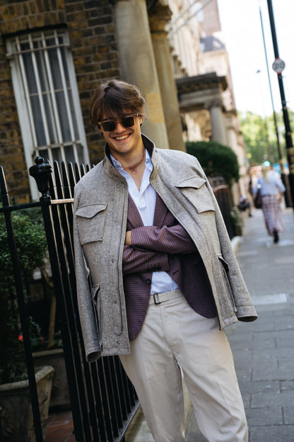 A man stands on a city sidewalk, smiling. He wears sunglasses, a grey jacket draped over a purple blazer, and a white shirt. His look is completed with light trousers and the Herno Beige Melange Loro Piana Dream Tweed Field Coat. Behind him are iron railings and brick buildings, with greenery and people visible in the background. , available at Richard Gelding Mayfair