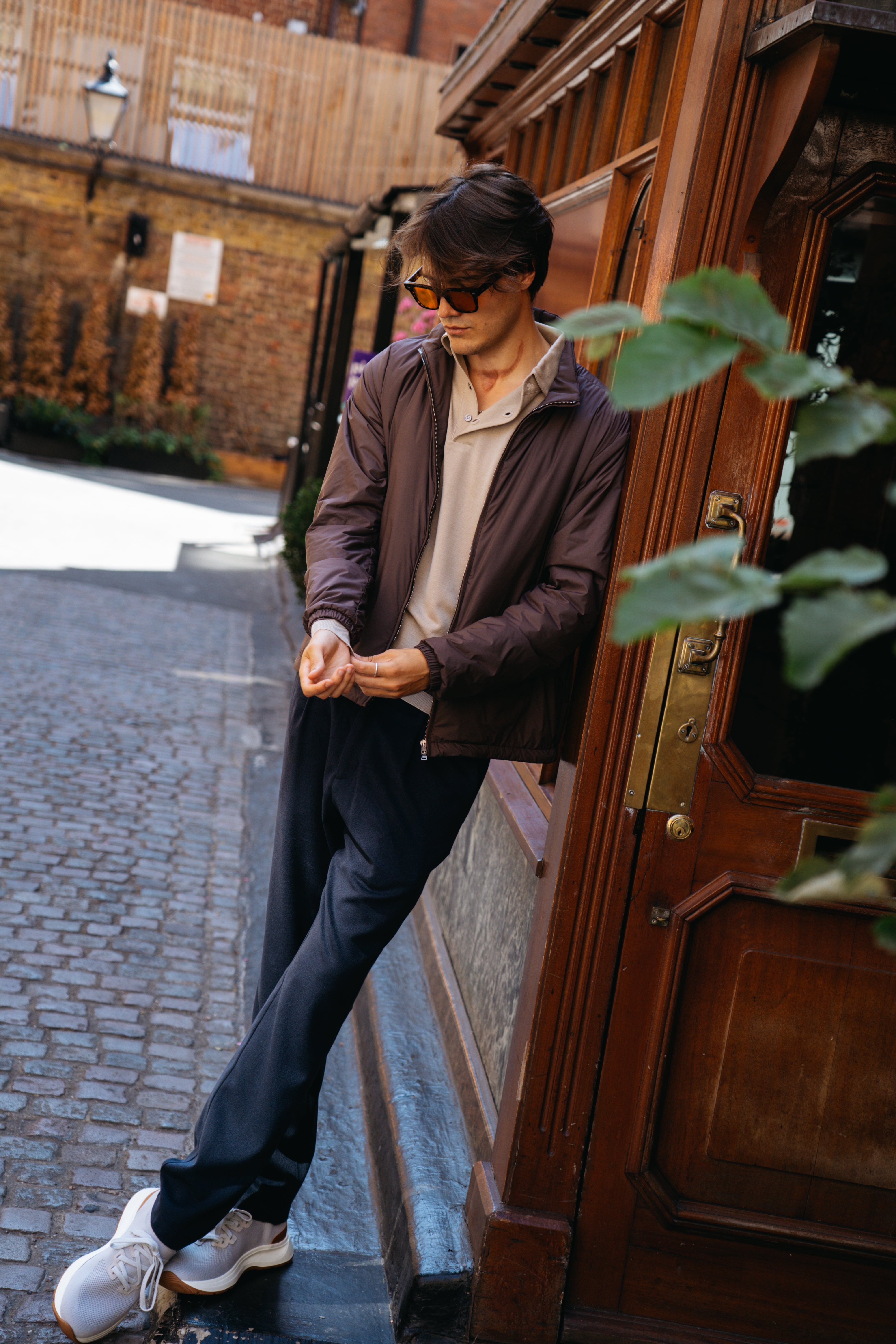 A man in sunglasses, a brown jacket, beige polo, dark pants, and white sneakers stands casually against a wooden doorway on a cobblestone street, adjusting his sleeve. Sunlight highlights him as green leaves partially frame the scene. His tailored look is completed with Herno’s Navy Blue Wool/Modal Melange Flannel Drawstring Resort Trousers—expertly crafted in Italy—embodying an effortless sense of style. , available at Richard Gelding Mayfair