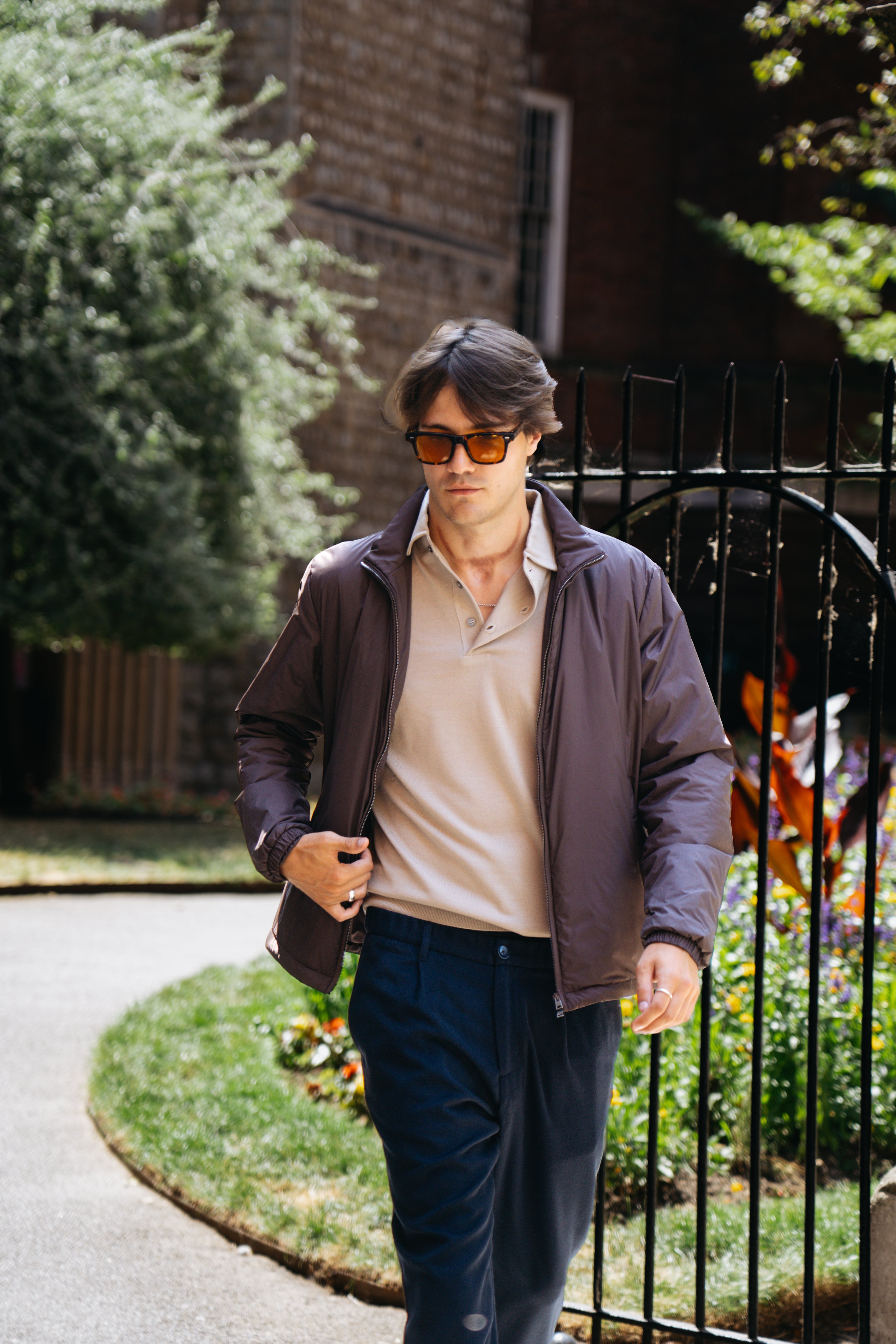 A young man walks outdoors near a black iron fence. He wears amber sunglasses, a brown bomber jacket, a beige polo shirt, and Herno Navy Blue Wool/Modal Melange Flannel Drawstring Resort Trousers, made in Italy. Green trees and a brick building are visible in the background. , available at Richard Gelding Mayfair