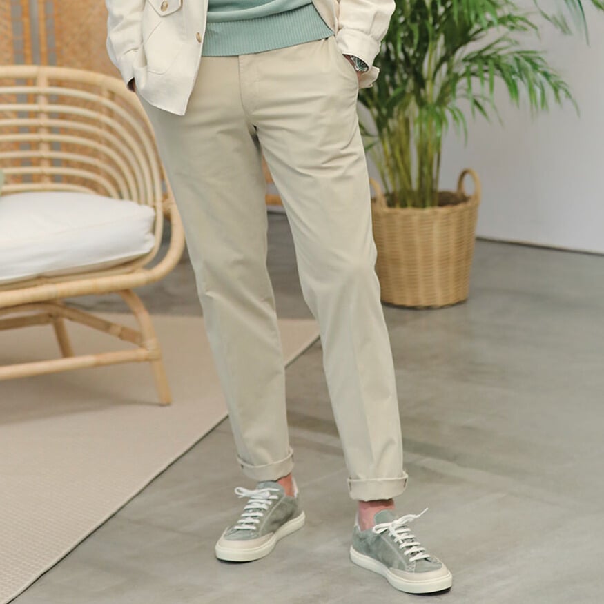 A person wears Hiltl Khaki Cotton Stretch Regular Fit Chinos, a light jacket, and green sneakers, standing on a gray floor beside a rattan chair and potted plant.