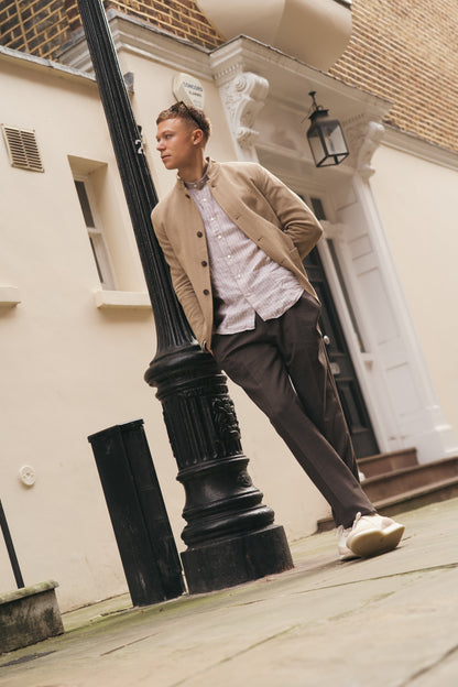 A man in a beige jacket, dark trousers, and an Alessandro Gherardi Brown Stripe Linen Mandarin Collar Shirt leans against a black lamppost on a stone-paved street before cream buildings with white trim and steps, hands in his pockets. , available at Richard Gelding Mayfair