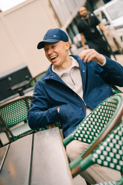 A smiling man sits outside at a café table wearing the Paige Dove Grey Ribbed Cotton Quarter Zip, paired with a blue cap and jacket. He is turned to the side, arm resting on a green-and-white chair. Another person and a white vehicle are blurred in the background. , available at Richard Gelding Mayfair