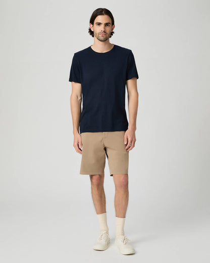 A man with medium-length dark hair stands facing forward against a light gray background. He wears a plain navy T-shirt, Paige Beige Farro Phillips Cotton Stretch Shorts, white crew socks, and white sneakers. His expression is neutral. , available at Richard Gelding Mayfair