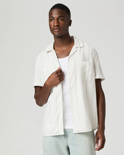 A man with short hair and a neutral expression stands against a plain light background wearing the Paige Ivory Stripe Cotton Blend 'Jacobson' Camp Collar Shirt over a white tank top and light blue jeans, facing slightly right with one hand near his collar. , available at Richard Gelding Mayfair