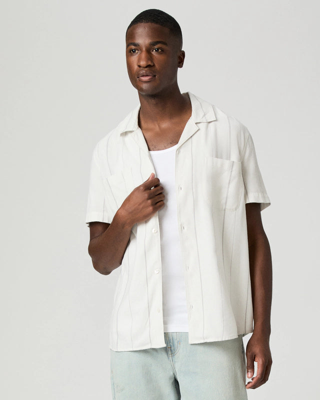 A man with short hair and a neutral expression stands against a plain light background wearing the Paige Ivory Stripe Cotton Blend 'Jacobson' Camp Collar Shirt over a white tank top and light blue jeans, facing slightly right with one hand near his collar. , available at Richard Gelding Mayfair