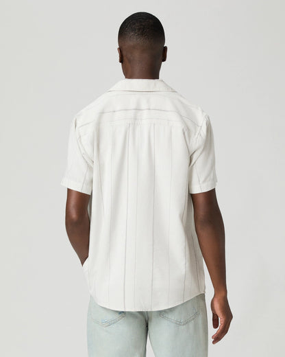 A man faces away from the camera in Paige's Ivory Stripe Cotton Blend 'Jacobson' Camp Collar Shirt, featuring subtle vertical stripes and short sleeves, paired with light blue jeans. A light gray background emphasizes his relaxed pose. , available at Richard Gelding Mayfair