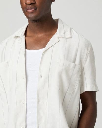 A person with medium skin tone wears the Paige Ivory Stripe Cotton Blend 'Jacobson' Camp Collar Shirt over a white ribbed tank, showing only their upper torso against a plain light gray background and a neutral expression. , available at Richard Gelding Mayfair