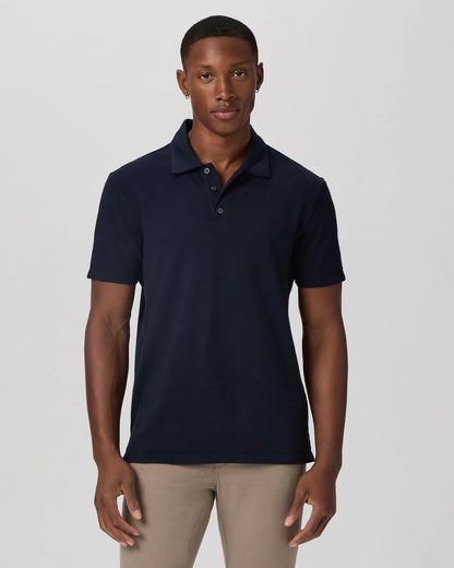 A man with short hair stands facing forward, arms relaxed, wearing the Paige Navy Pique Cotton Demille Short Sleeve Polo and light beige pants against a plain light gray background, his expression neutral. , available at Richard Gelding Mayfair