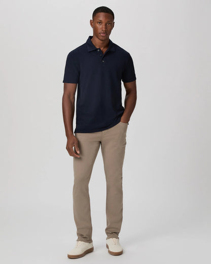 A man stands against a white background, wearing the Paige Navy Pique Cotton Demille Short Sleeve Polo, light khaki pants, and white sneakers. He has a neutral expression, with his left hand in his pocket and right arm at his side. , available at Richard Gelding Mayfair