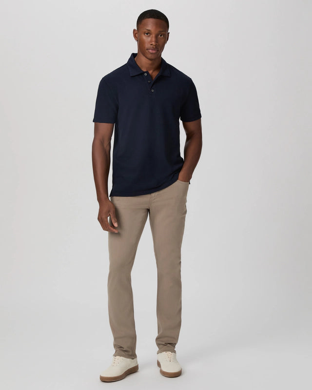 A man stands against a white background, wearing the Paige Navy Pique Cotton Demille Short Sleeve Polo, light khaki pants, and white sneakers. He has a neutral expression, with his left hand in his pocket and right arm at his side. , available at Richard Gelding Mayfair