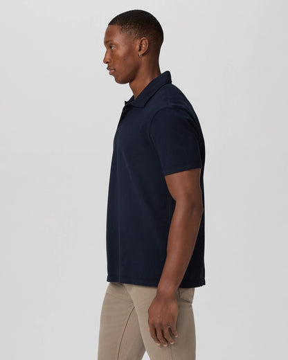 A man stands in profile facing left, wearing the Paige Navy Pique Cotton Demille Short Sleeve Polo and light beige pants. The plain, light background highlights the shirt’s fit and style. , available at Richard Gelding Mayfair