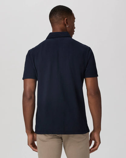 A man with short hair stands with his back to the camera, wearing the Paige Navy Pique Cotton Demille Short Sleeve Polo and light khaki pants, his arms relaxed at his sides against a plain light gray background. , available at Richard Gelding Mayfair