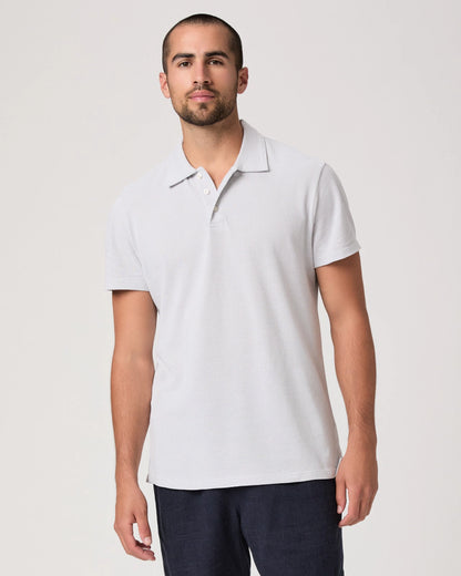 A man with short hair and a trimmed beard stands facing forward in the Paige Bristol Blue Demille Cotton Polo, a classic short-sleeve men's shirt, paired with dark pants. He has a neutral expression against an off-white background. , available at Richard Gelding Mayfair