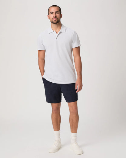 A man with short hair and a beard stands against a plain light background, wearing the Paige Bristol Blue Demille Cotton Polo, dark navy shorts, white crew socks, and white sneakers. One hand is in his pocket as he looks towards the camera. , available at Richard Gelding Mayfair