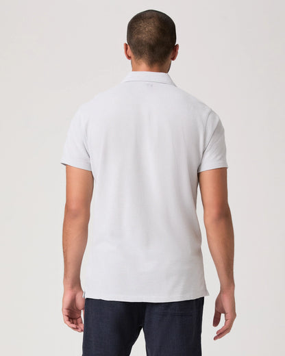 A man with short hair is seen from behind wearing the Paige Bristol Blue Demille Cotton Polo and dark pants, standing against a plain, light background with his arms relaxed at his sides. , available at Richard Gelding Mayfair