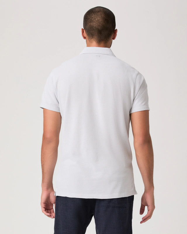 A man with short hair is seen from behind wearing the Paige Bristol Blue Demille Cotton Polo and dark pants, standing against a plain, light background with his arms relaxed at his sides. , available at Richard Gelding Mayfair