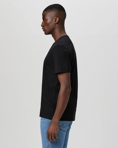 A young man in profile faces left, wearing a Paige Black Cotton Sorden Crew Neck T-Shirt with light blue jeans. Set against a solid light gray background, his neutral expression highlights the simplicity and casual style of the outfit. , available at Richard Gelding Mayfair