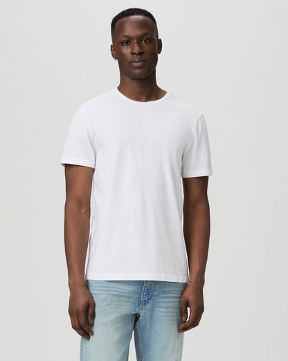 A man stands facing forward against a light gray background, wearing a Paige White Cotton Sorden Crew Neck T-Shirt and light blue jeans. His expression is neutral, and his arms rest naturally at his sides. , available at Richard Gelding Mayfair