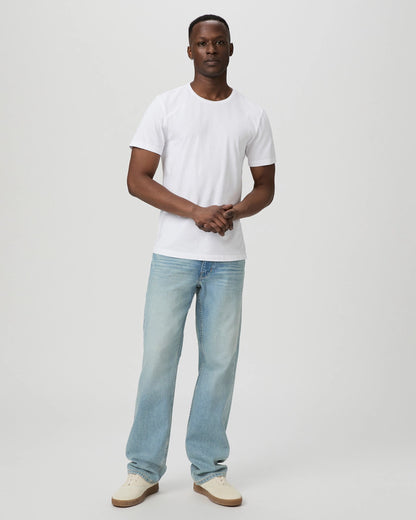 A man stands against a plain light gray background, wearing the Paige White Cotton Sorden Crew Neck T-Shirt, paired with light blue jeans and beige sneakers. He looks forward with a neutral expression, hands loosely clasped in front. , available at Richard Gelding Mayfair