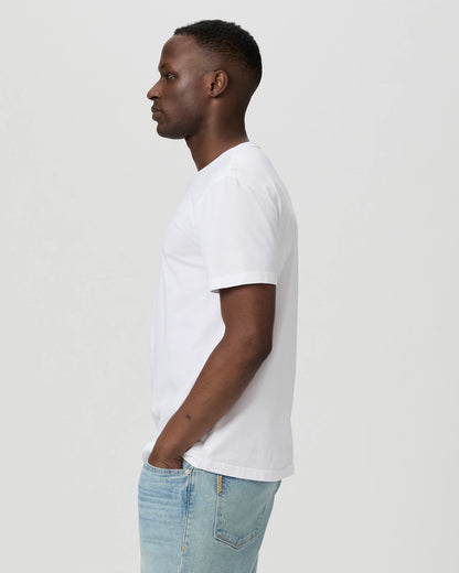 A man stands in profile facing left, wearing a Paige White Cotton Sorden Crew Neck T-Shirt and light blue jeans with his hands in his pockets against a plain light gray background. , available at Richard Gelding Mayfair