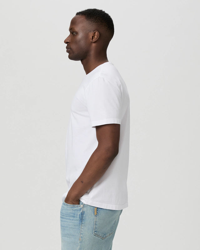 A man stands in profile facing left, wearing a Paige White Cotton Sorden Crew Neck T-Shirt and light blue jeans with his hands in his pockets against a plain light gray background. , available at Richard Gelding Mayfair