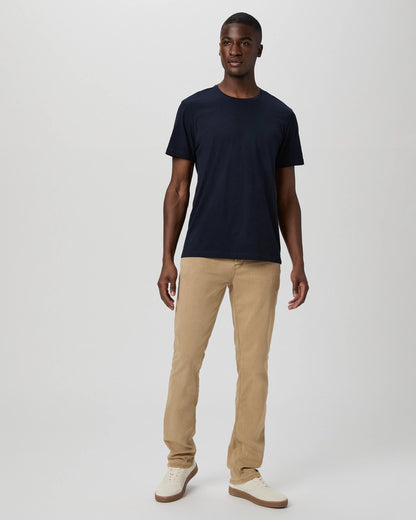 A man stands against a light gray background, wearing the Paige Deep Anchor Navy Cotton Sorden Crew Neck, beige straight-leg pants, and white sneakers with tan soles. His arms are relaxed at his sides and he has a neutral expression. , available at Richard Gelding Mayfair