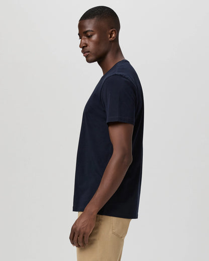 A man stands in profile against a light gray background, wearing Paige's Deep Anchor Navy Cotton Sorden Crew Neck and light tan pants. His arms rest by his sides as he gazes slightly downward with a neutral expression. , available at Richard Gelding Mayfair