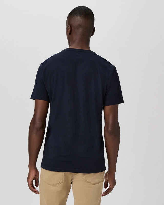 A person faces away from the camera, wearing the Paige Deep Anchor Navy Cotton Sorden Crew Neck t-shirt with light beige pants. The background is light gray, uncluttered; their left arm hangs relaxed and right arm is slightly bent. , available at Richard Gelding Mayfair