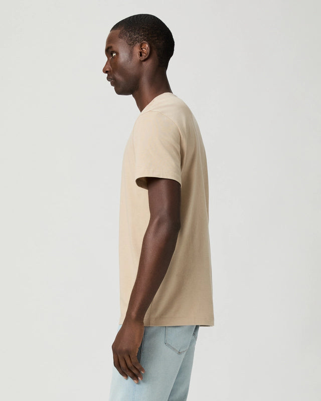 A man in profile faces left against a plain light background, wearing the Paige Natural Beige Cotton Sorden Crew Neck T-Shirt and light blue jeans. His arms hang naturally at his sides, and his hair is short and closely cropped. , available at Richard Gelding Mayfair