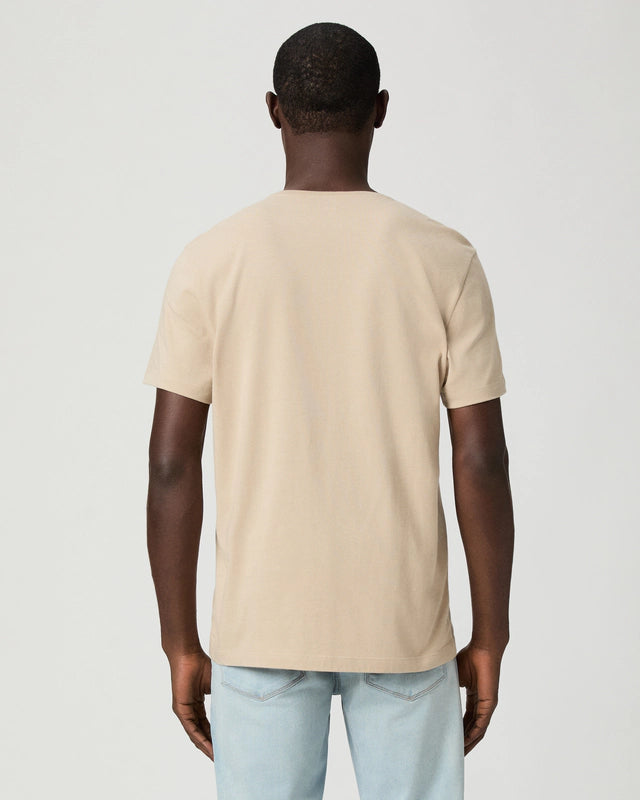 A man faces away from the camera, wearing a Paige Natural Beige Cotton Sorden Crew Neck T-Shirt and light blue jeans. He stands against a plain light gray background with his arms relaxed at his sides. , available at Richard Gelding Mayfair