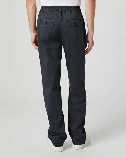A person stands facing away, wearing Paige Navy Cotton Stretch 'Eberton' Trousers featuring an elastic waistband and two back pockets with buttoned flaps. They are also in white shoes and a white shirt, against a plain light background. , available at Richard Gelding Mayfair