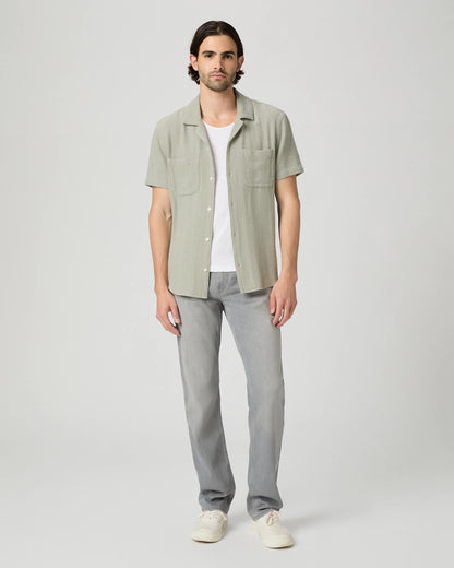 A man faces forward against a plain background, wearing Paige Grey Wainwright Normandie Regular Fit Jeans, a light green short-sleeve shirt over a white t-shirt, and white sneakers. He has medium brown hair and rests his arms by his sides. , available at Richard Gelding Mayfair