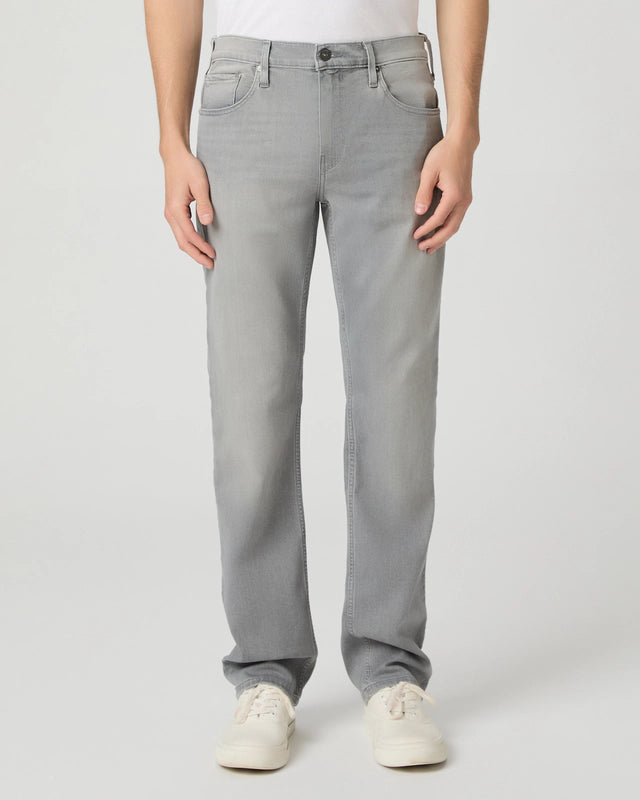 A person stands facing forward, wearing Paige Grey Wainwright Normandie Regular Fit Jeans with a white tucked-in shirt and white sneakers. Only the lower body is shown, hands at the sides, against a plain light grey background. , available at Richard Gelding Mayfair